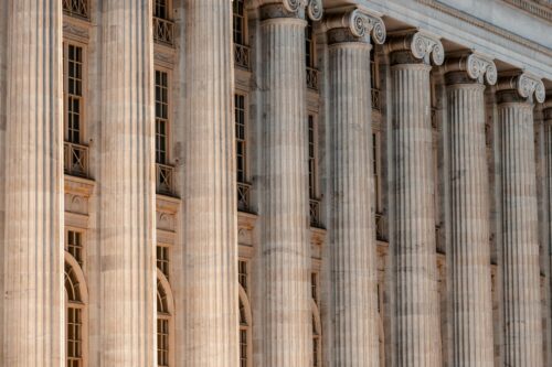 Rows of grand, classical stone columns with ornate capitals line a historic building facade, exuding a sense of strength and elegance in warm light. https://unsplash.com/photos/a-large-building-with-columns-and-a-clock-on-the-front-of-it-_JEiyOfC2y8?utm_source=unsplash&utm_medium=referral&utm_content=creditShareLink