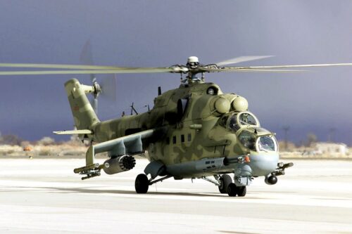 A military helicopter with camouflage paint rests on the runway under a cloudy sky. The rotors are slightly blurred, indicating recent motion.