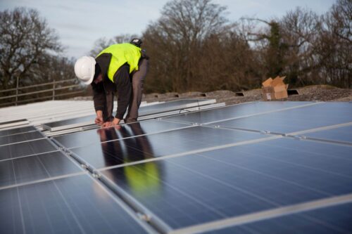 A worker in a yellow vest and hard hat installing solar panels. The setting is outdoors with trees in the background under a cloudy sky. Photo by Quang Nguyen Vinh: https://www.pexels.com/photo/expansive-solar-farm-in-vietnam-s-binh-thuan-province-35105443/