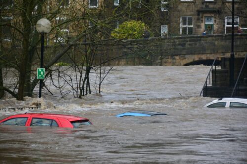 Floodwaters submerge cars on a street beside a riverbank. Trees and a streetlight are partially submerged. The scene conveys urgency and chaos. https://unsplash.com/photos/car-on-body-of-water-4zxp5vlmvnI?utm_source=unsplash&utm_medium=referral&utm_content=creditShareLink