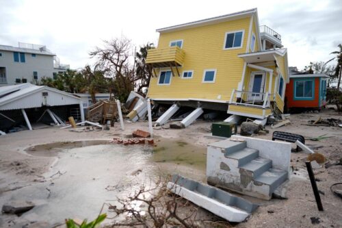 A yellow house leans precariously on damaged stilts after a storm, surrounded by debris and muddy water, evoking a sense of devastation and chaos.
