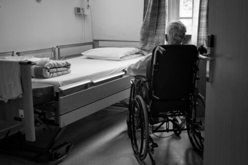 A person in a wheelchair sits beside a neatly made hospital bed, gazing out the window. The scene is calm and contemplative, with soft light filtering in. https://unsplash.com/photos/a-person-in-a-wheelchair-in-a-room-fUBZHfDAVmc?utm_source=unsplash&utm_medium=referral&utm_content=creditShareLink