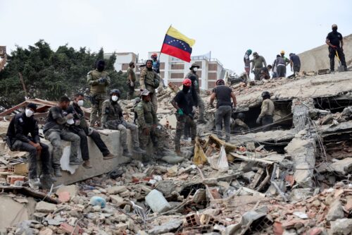 CARABELLADA, VENEZUELA - JUNE 28: Rescuers search for victims at a collapsed building as one raises a Venezuelan flag following a magnitude 7.2 earthquake that struck Venezuela and other regions in the Caribbean, on June 28, 2026 in Carabellada, La Guaira, Venezuela. According to the U.S. Geological Survey (USGS), the main earthquake on June 24, 2026 was followed by a 7.5-magnitude aftershock less than a minute later. The number of fatal victims increased to more than 1000, while the number of injured people exceeded 3,000. More than 70,000 people are reported missing. (Photo by Jesus Vargas/Getty Images)