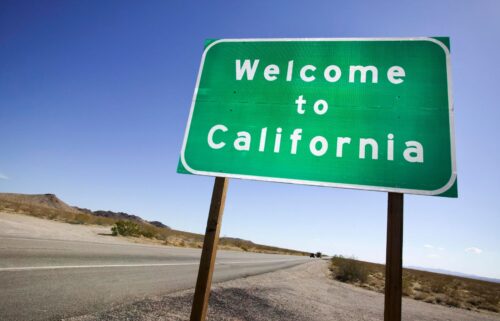 A green rectangular road sign with white text reading Welcome to California, mounted on wooden posts beside a desert highway under a clear blue sky.