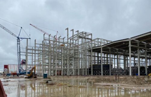 Construction site with steel framework and cranes under a cloudy sky. Muddy ground with puddles reflects the industrial structure, conveying stalled progress.