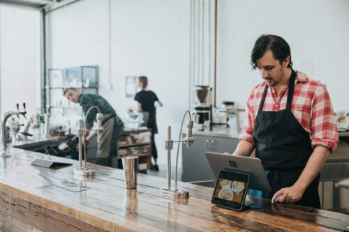 Barista in a red plaid shirt and apron uses a tablet at a wooden counter in a modern café; another worker busy in the background, creating a focused and industrious atmosphere. https://unsplash.com/photos/people-in-kitchen-X_bbCAYzlTs?utm_source=unsplash&utm_medium=referral&utm_content=creditShareLink